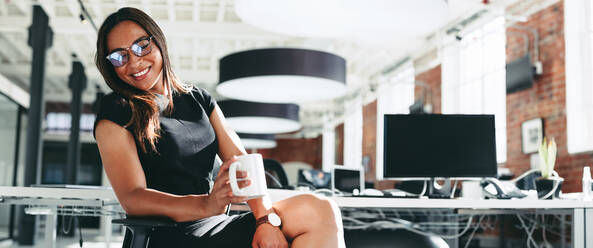 Glückliche junge Geschäftsfrau, die fröhlich lächelt, während sie eine Tasse Kaffee in der Hand hält. Junge Geschäftsfrau, die allein an einem modernen Arbeitsplatz sitzt. - JLPSF07928