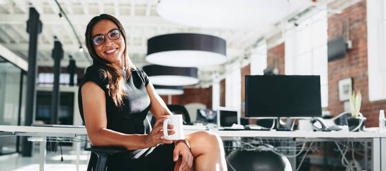 Businesswoman having a coffee break at work. Happy young businesswoman smiling cheerfully while holding a cup of coffee. Young businesswoman sitting alone in a modern workplace. - JLPSF07929
