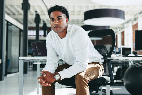 Digital developer looking at the camera in a modern workplace. Confident young businessman sitting alone in an office. Creative businessman taking a break while working on a new project. - JLPSF07945