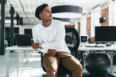 Thinking of new software ideas. Creative businessman looking away thoughtfully while sitting alone in a modern workplace. Young software developer taking a break while working in an office. - JLPSF07946