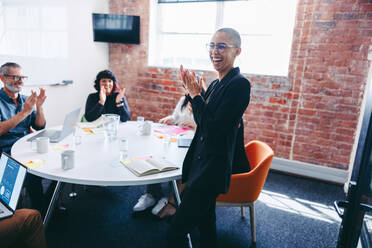 Junge Geschäftsfrau, die von ihrem Team während einer Besprechung in einem modernen Büro beklatscht wird. Erfolgreiche junge Geschäftsfrau, die fröhlich lächelt, während sie von ihren Kollegen in einem Besprechungsraum gelobt wird. - JLPSF07987