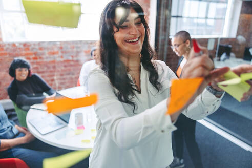 Lächelnde Geschäftsfrau, die Haftnotizen an eine Glaswand klebt, mit ihren Kollegen im Hintergrund. Konzentrierte junge Geschäftsfrau, die ihre Ideen mit ihrem Team an einem modernen Arbeitsplatz teilt. - JLPSF07990
