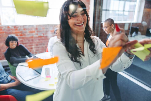 Smiling businesswoman sticking adhesive notes to a glass wall with her colleagues in the background. Focused young businesswoman sharing her ideas with her team in a modern workplace. - JLPSF07990