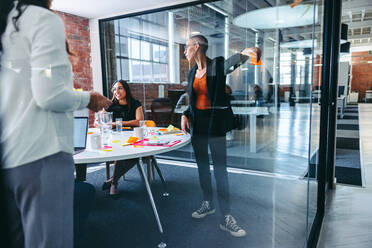 Geschäftsleute beim Brainstorming in einem Büro. Zwei kreative Geschäftsfrauen stehen vor einer Glaswand, während ihre Kollegen im Hintergrund sitzen. Geschäftsfrauen teilen ihre Ideen mit ihrem Team. - JLPSF07993