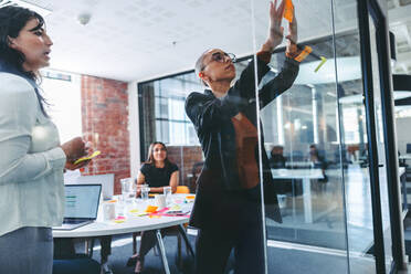 Kreative Geschäftsfrauen, die Haftnotizen an eine Glaswand kleben, mit ihren Kollegen im Hintergrund. Zwei konzentrierte Geschäftsfrauen, die ihre Ideen mit ihrem Team in einem modernen Büro austauschen. - JLPSF07994