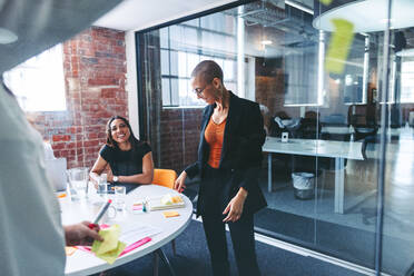 Kreative Geschäftsfrauen tauschen ihre Ideen in einem modernen Büro aus. Zwei konzentrierte junge Geschäftsfrauen kleben Haftnotizen an eine Glaswand, während sie mit ihren Kollegen ein Brainstorming durchführen. - JLPSF07996