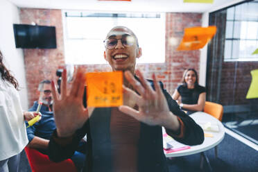Smiling businesswoman sticking adhesive notes to a glass wall with her colleagues in the background. Happy young businesswoman sharing her ideas with her team in a modern office. - JLPSF07997