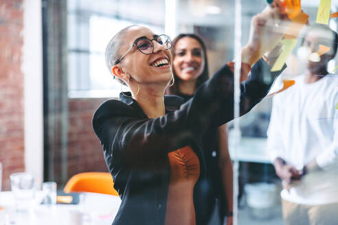 Lächelnde Geschäftsfrau, die ihre Ideen mit ihren Kollegen an einem kreativen Arbeitsplatz teilt. Selbstbewusste junge Geschäftsfrau, die Haftnotizen an eine Glaswand klebt, während ihr Team im Hintergrund steht. - JLPSF08000