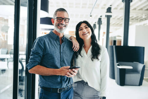 Fröhlich lächelnde Kollegen in einem Büro. Glücklicher reifer Geschäftsmann, der ein Smartphone hält, während er mit seiner Kollegin an einem modernen Arbeitsplatz steht. Geschäftsleute, die als Team arbeiten. - JLPSF08003