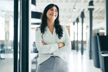 Cheerful businesswoman standing with her arms crossed. Happy young businesswoman looking away with a smile on her face in a modern workplace. Successful businesswoman standing alone in an office. - JLPSF08006