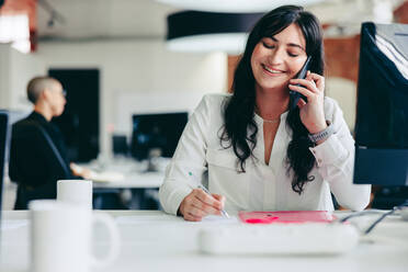 Glückliche Geschäftsfrau, die Geschäftspläne macht, während sie ein Telefongespräch führt. Kreative Geschäftsfrau, die sich mit ihren Geschäftskunden in einem modernen Büro trifft. Junge Geschäftsfrau, die fröhlich lächelt. - JLPSF08010