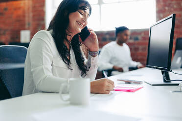 Lächelnde Geschäftsfrau, die einen Anruf im Büro entgegennimmt. Kreative Geschäftsfrau, die in einem modernen Büro sitzend mit ihren Geschäftskunden spricht. Junge Geschäftsfrau, die Geschäftspläne macht. - JLPSF08011