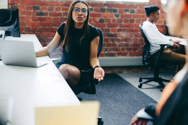 Zusammenarbeit im Büro: Junge Geschäftsfrau, die sich am Schreibtisch mit ihrem Kollegen unterhält. Kreative Geschäftsfrau, die an einem modernen Arbeitsplatz Ideen mit ihrer Kollegin austauscht. - JLPSF08012