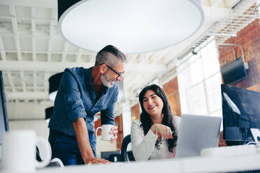 Successful businesspeople laughing together while working on a new project in an office. Happy mature businessman collaborating with his female colleague in a modern workplace. - JLPSF08014