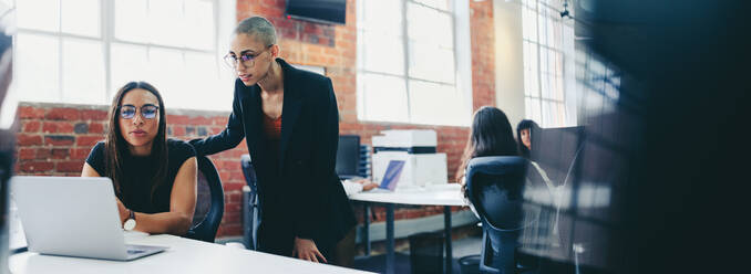 Zwei junge Geschäftsfrauen, die in einem kreativen Büro zusammenarbeiten. Weibliche Kollegen, die gemeinsam einen Laptop benutzen, während sie an einem neuen Projekt arbeiten. Geschäftsfrauen, die zusammenarbeiten und Ideen austauschen. - JLPSF08020