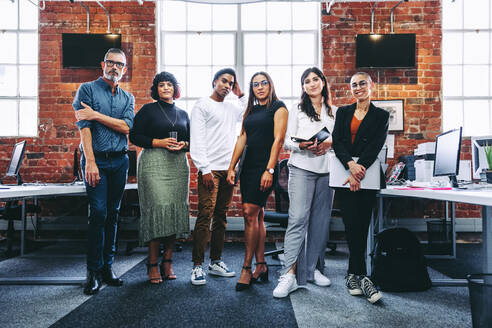 Ein buntes Team von Geschäftsleuten, die in einem Büro zusammenstehen. Eine Gruppe von modernen Geschäftsleuten, die an einem kreativen Arbeitsplatz in die Kamera schauen. Geschäftskollegen, die fröhlich aussehen. - JLPSF08029