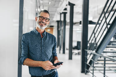 Happy mature businessman holding a smartphone and smiling at the camera. Creative businessman standing alone in a modern workplace. Experienced businessman communicating with his clients. - JLPSF08033