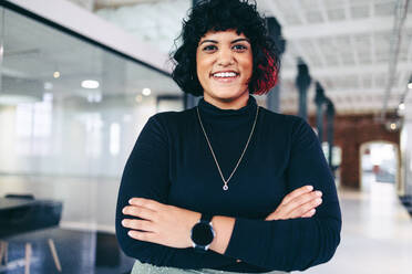 Female entrepreneur smiling in a creative office. Happy mid-adult businesswoman looking at the camera while standing with her arms crossed. Successful businesswoman standing alone in a modern workplace. - JLPSF08041