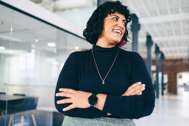 Female company owner looking away with a cheerful smile. Happy mid-adult businesswoman standing with her arms crossed in a creative office. Successful businesswoman standing alone in a modern workplace. - JLPSF08042