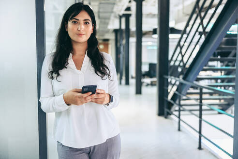 Intelligente Geschäftsfrau, die ein Smartphone in einem Büro hält. Selbstbewusste Geschäftsfrau, die in die Kamera schaut, während sie allein an einem modernen Arbeitsplatz steht. Mid-Adult-Geschäftsfrau, die mit ihren Kunden kommuniziert. - JLPSF08045