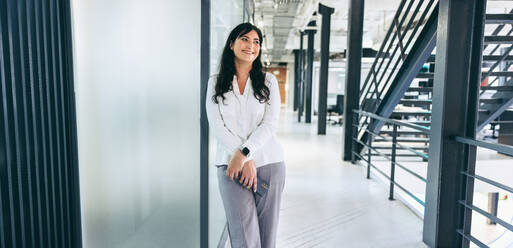 Businesswoman smiling while standing in a creative workplace. Happy mid-adult businesswoman holding a smartphone in a modern office. Successful businesswoman looking cheerful. - JLPSF08046