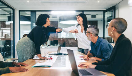 Two cheerful businesswomen elbow bumping each other before a meeting in a boardroom. Group of businesspeople attending a briefing in a modern workplace. Businesspeople taking COVID-19 precautions. - JLPSF08092