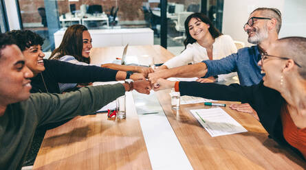 Fröhliche Gruppe von Geschäftsleuten, die ihre Fäuste zusammenbringen, während sie in einem Sitzungssaal sitzen. Unterschiedliche Gruppe von Kollegen, die während einer Sitzung an einem modernen Arbeitsplatz fröhlich lächeln. - JLPSF08100