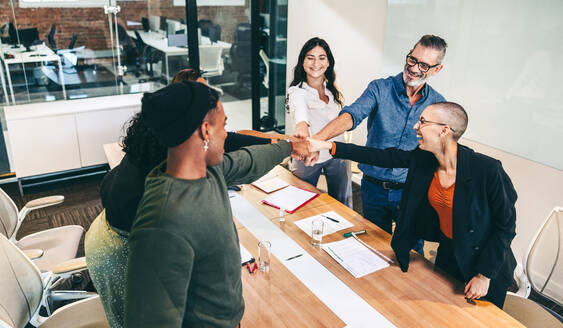 Confident group of businesspeople bringing their fists together while standing in a boardroom. Diverse group of colleagues smiling cheerfully during a meeting in modern workplace. - JLPSF08102