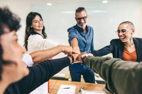 Happy group of businesspeople bringing their fists together while standing in a boardroom. Diverse group of businesspeople smiling cheerfully while standing together in a huddle. - JLPSF08103