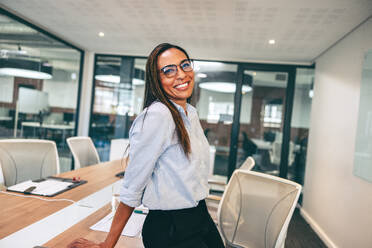 Fröhlich lächelnde Geschäftsfrau in einem Büro. Fröhliche Geschäftsfrau, die sich in einem Sitzungssaal an einen Tisch lehnt. Fröhliche Unternehmerin in legerer Businesskleidung an einem kreativen Arbeitsplatz. - JLPSF08106