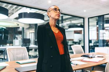 Selbstbewusste junge Geschäftsfrau, die nachdenklich wegschaut, während sie allein in einem Sitzungssaal steht. Junge Unternehmerin in legerer Businesskleidung an einem modernen Arbeitsplatz. - JLPSF08108