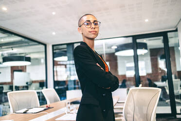 Selbstbewusste Unternehmerin, die in einem Büro in die Kamera schaut. Kreative junge Geschäftsfrau, die mit verschränkten Armen daneben steht. Junge Geschäftsfrau in Business Casual in einem modernen Büro. - JLPSF08111