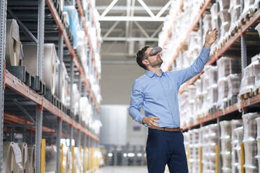 Businessman wearing virtual reality simulator gesturing in warehouse - JOSEF14192