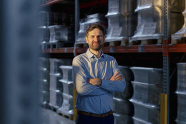 Smiling businessman with arms crossed standing by rack in warehouse - JOSEF14210