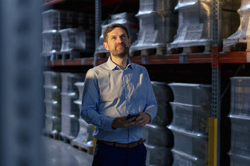 Thoughtful businessman with smart phone standing by rack in warehouse - JOSEF14211