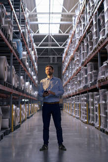 Businessman with smart phone standing amidst racks in warehouse - JOSEF14212