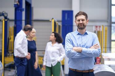 Ein lächelnder Geschäftsmann steht mit Kollegen im Hintergrund in einer Fabrik - JOSEF14221