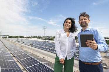 Smiling businessman holding tablet PC standing by businesswoman in front of solar panels - JOSEF14343