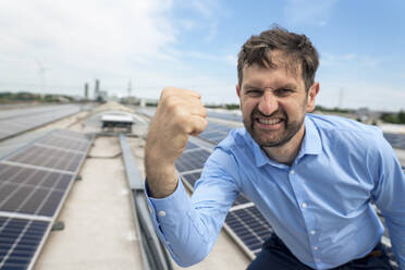 Happy businessman gesturing fist in front of solar panels - JOSEF14361