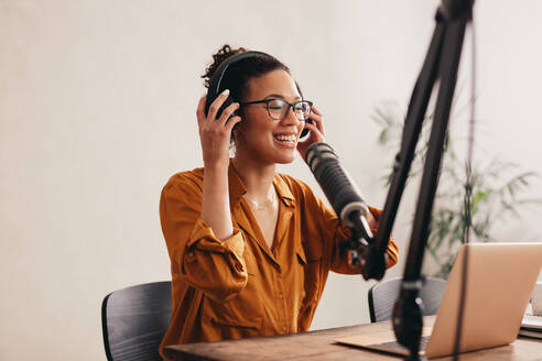 Eine Frau mit Kopfhörern bereitet sich auf die Aufnahme eines Podcasts im Heimstudio vor. Eine Frau arbeitet von zu Hause aus mit einem Laptop und einem Mikrofon auf dem Tisch. - JLPSF08128
