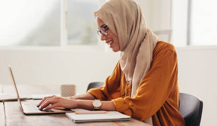 Woman wearing hijab working on laptop at home. Woman working from home using laptop and smiling. - JLPSF08145