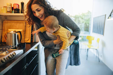 Baby boy wants to pick a cup from kitchen counter while in arms of his mother. Woman carrying his son reaching out to a cup on kitchen counter. - JLPSF08155