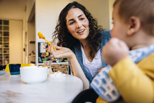 Eine Frau spricht mit ihrem Baby, während sie es zu Hause füttert. Eine fürsorgliche Mutter füttert ihr Kleinkind während ihres Mutterschaftsurlaubs. - JLPSF08161
