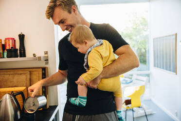 Vater trägt sein Baby und bereitet Kaffee in der Küche zu. Mann im Vaterschaftsurlaub mit seinem Kleinkind steht in der Küche und kocht Kaffee. - JLPSF08162