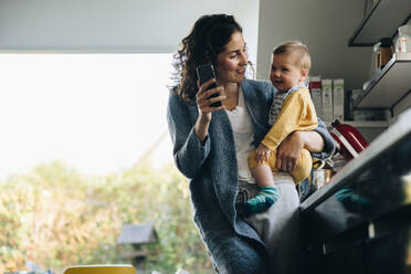 Frau zeigt ihrem Baby ihr Handy, während sie in der Küche steht. Mutter genießt ihre Zeit im Mutterschaftsurlaub mit ihrem Baby. - JLPSF08165