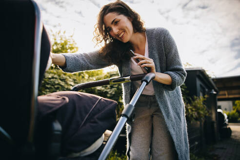 Fröhliche Mutter, die einen Kinderwagen schiebt und ihr Baby betrachtet. Glückliche Mutter mit Kinderwagen im Freien. - JLPSF08171