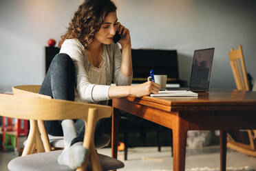 Relaxed woman sitting at table talking on mobile phone and taking notes. Woman handling her business while at home. - JLPSF08187