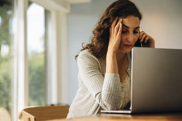 Woman looking stressed out while talking on a cellphone. Woman feeling stressed sitting at table with laptop and talking on mobile phone. - JLPSF08188