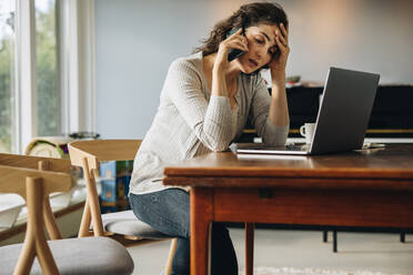 Woman having headache talking on cellphone while working from home. Woman sitting at table with laptop computer looking stressed out while talking over phone call. - JLPSF08189