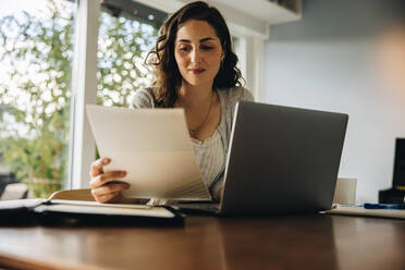Frau liest ein Dokument, während sie an einem Laptop arbeitet. Frau arbeitet von zu Hause aus. - JLPSF08191